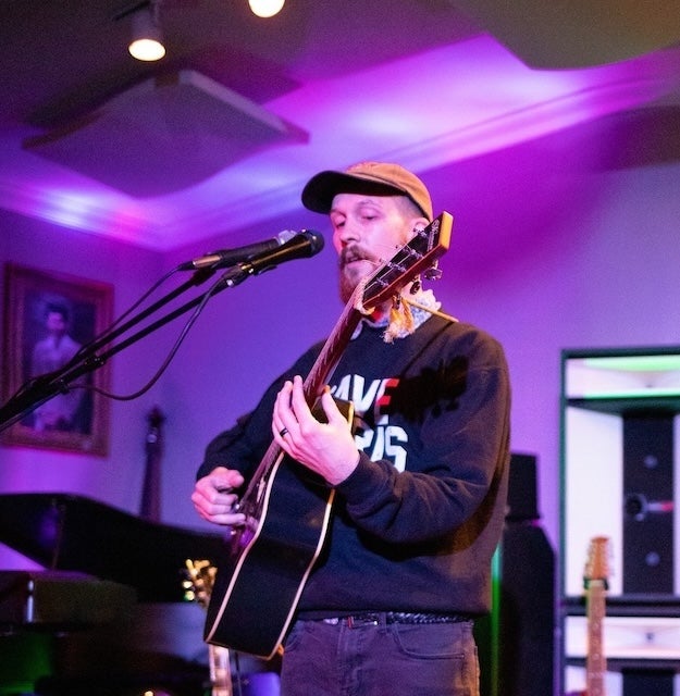 White man wearing black sweater and a brown baseball cap holding acoustic guitar and singing into a microphone