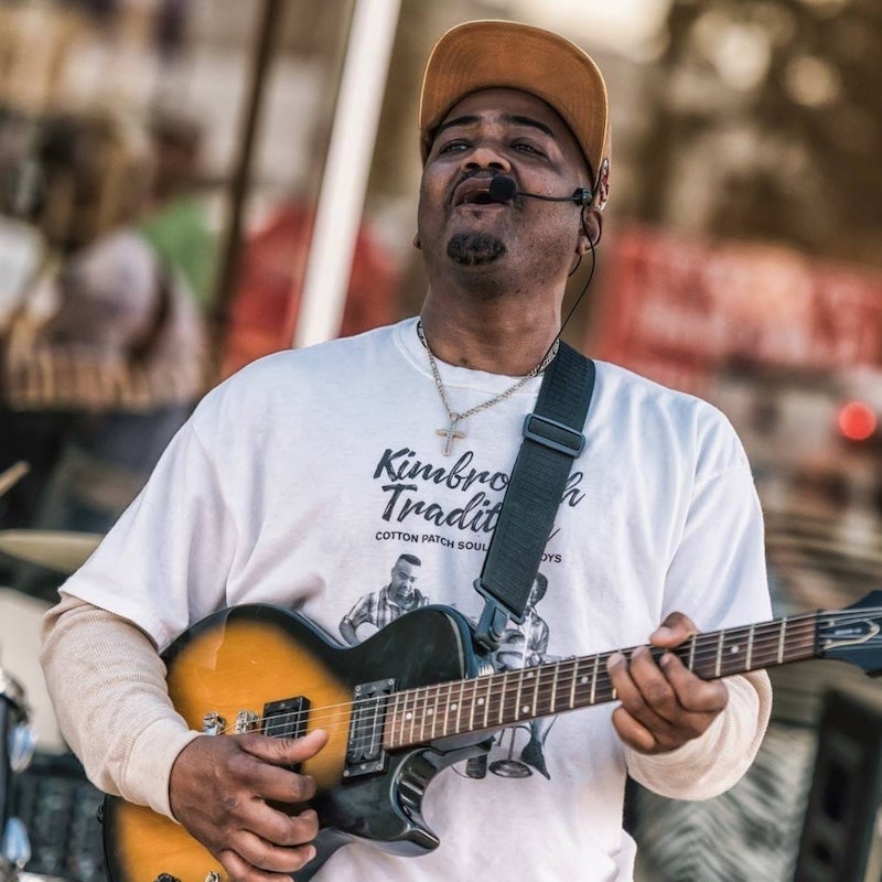 Middle aged black man wearing white T-shirt and brown baseball cap, holding electric guitar and singing into a headset microphone