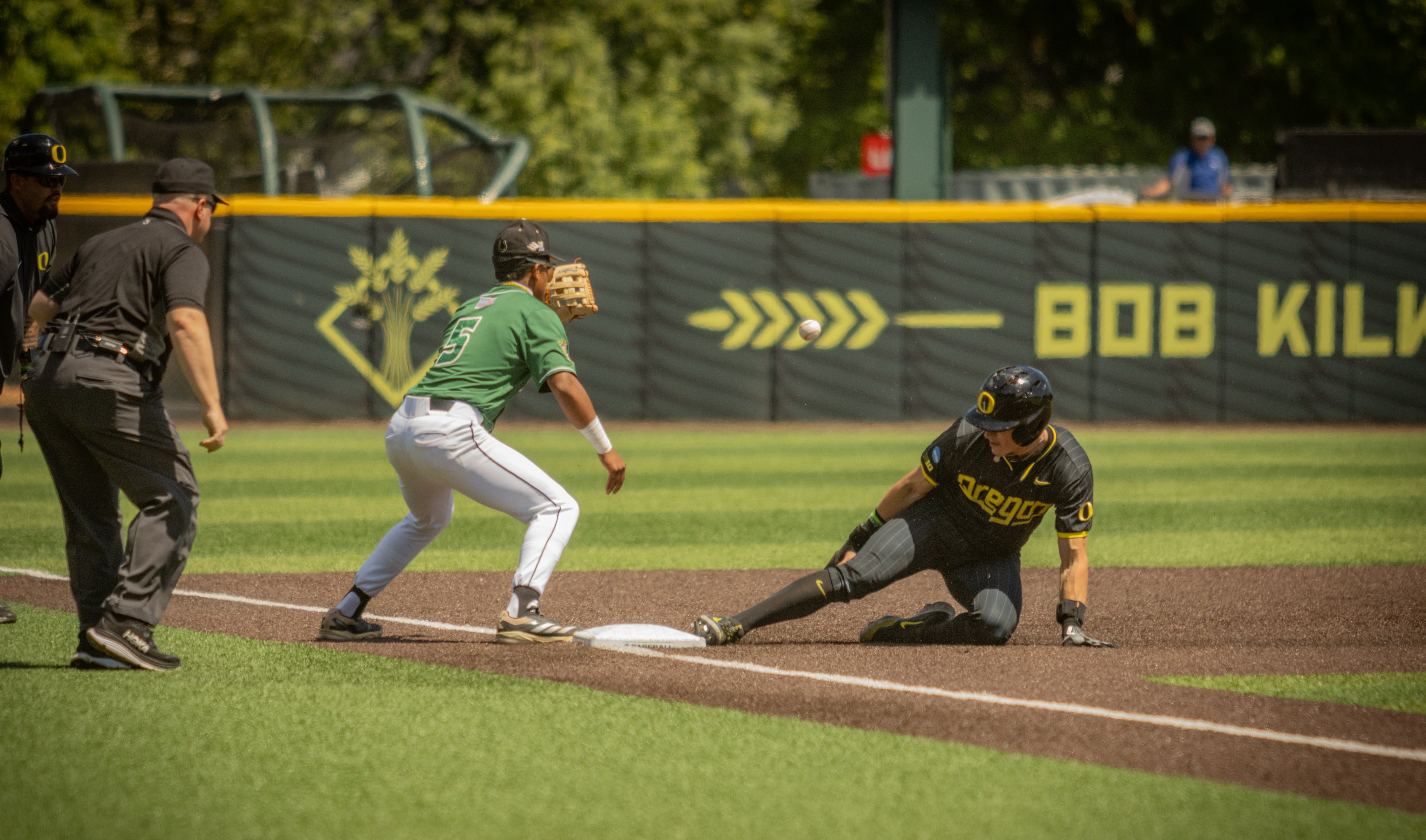 An Oregon baseball player slides in at base as Cal Poly attempts a throw for the out.