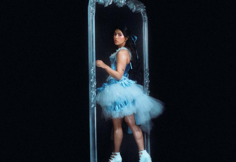 A black background with a woman in a blue dress and white boots walking into a mirror and turning to look at the camera 