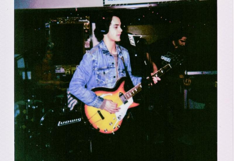 Polaroid photo reading "KWVA LIVE SESSIONS HENRY'S UNCLE 12-8-24" with young white man with black hair and denim jacket holding guitar singing into microphone